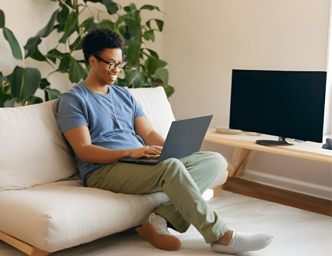 Sitting cross-legged on a plush couch, a man balances his laptop on his lap, the room filled with natural light from a nearby window.